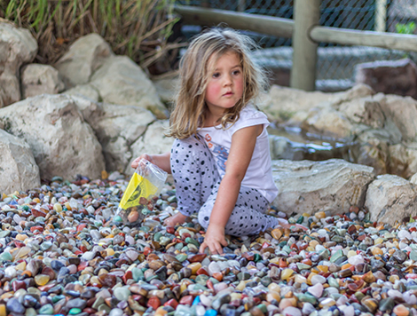 This picture shows a little girl sittingin a scrach patch of stones. There are hundreds of colourful stones underneath her.