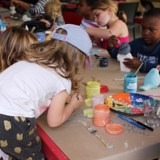 a bunch of children sitting at a table making crafts from clay.  There are a lot of open bottles of paint and in all sorts of colours