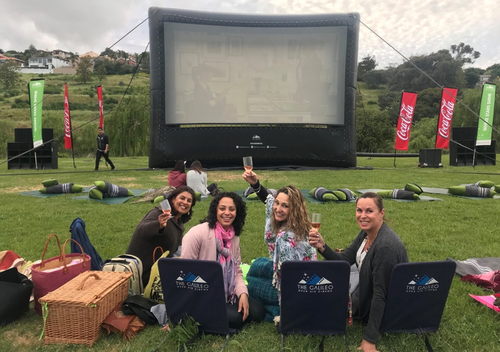 A group of woman toasting to their outdoor movie at the Galileo outdoor cinema. Theyre sitting on Galileo branded chairs in an open wine farm.
