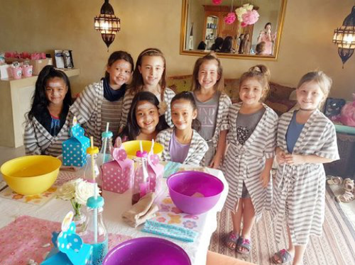 A group of little girls posing for a photo in front of their pamer table.  They&#x27;re all wearing gowns and have face masks on